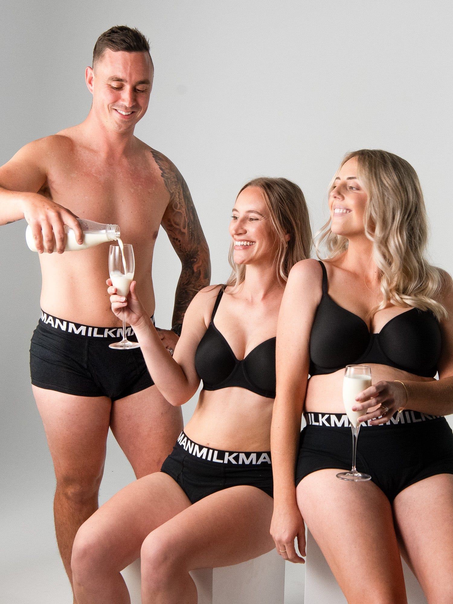 Two women in black underwear with &#39;Milkman&#39; branding, one pouring milk into a glass held by a man.